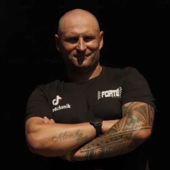 Mechanik w czarnej koszulce, z tatuażem na przedramieniu, stoi z założonymi rękami.